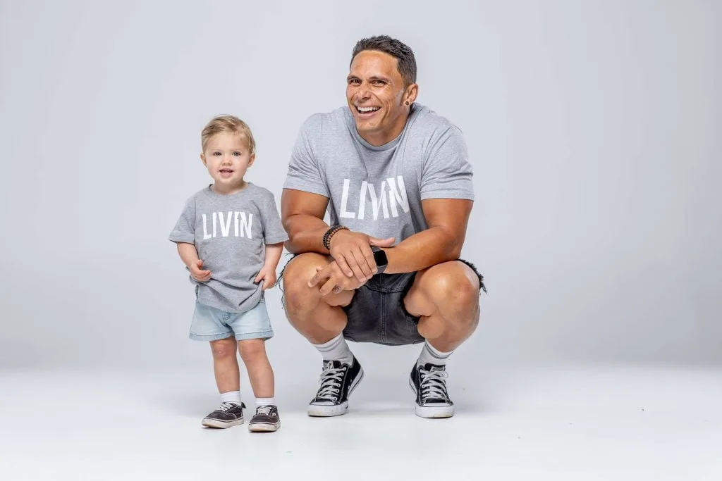 A man crouching beside a smiling toddler, both wearing grey LIVIN t-shirts